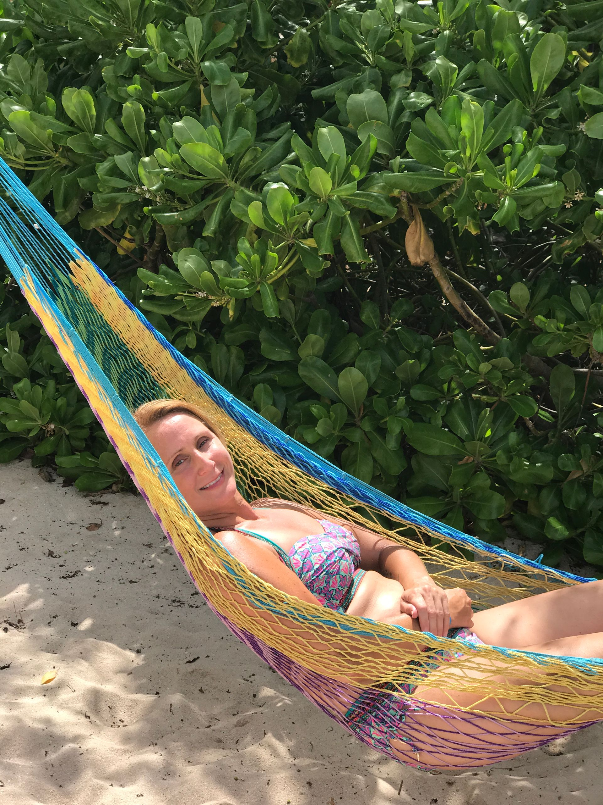 woman reclining in a colorful hammock beside dense green foliage on a sandy beach.