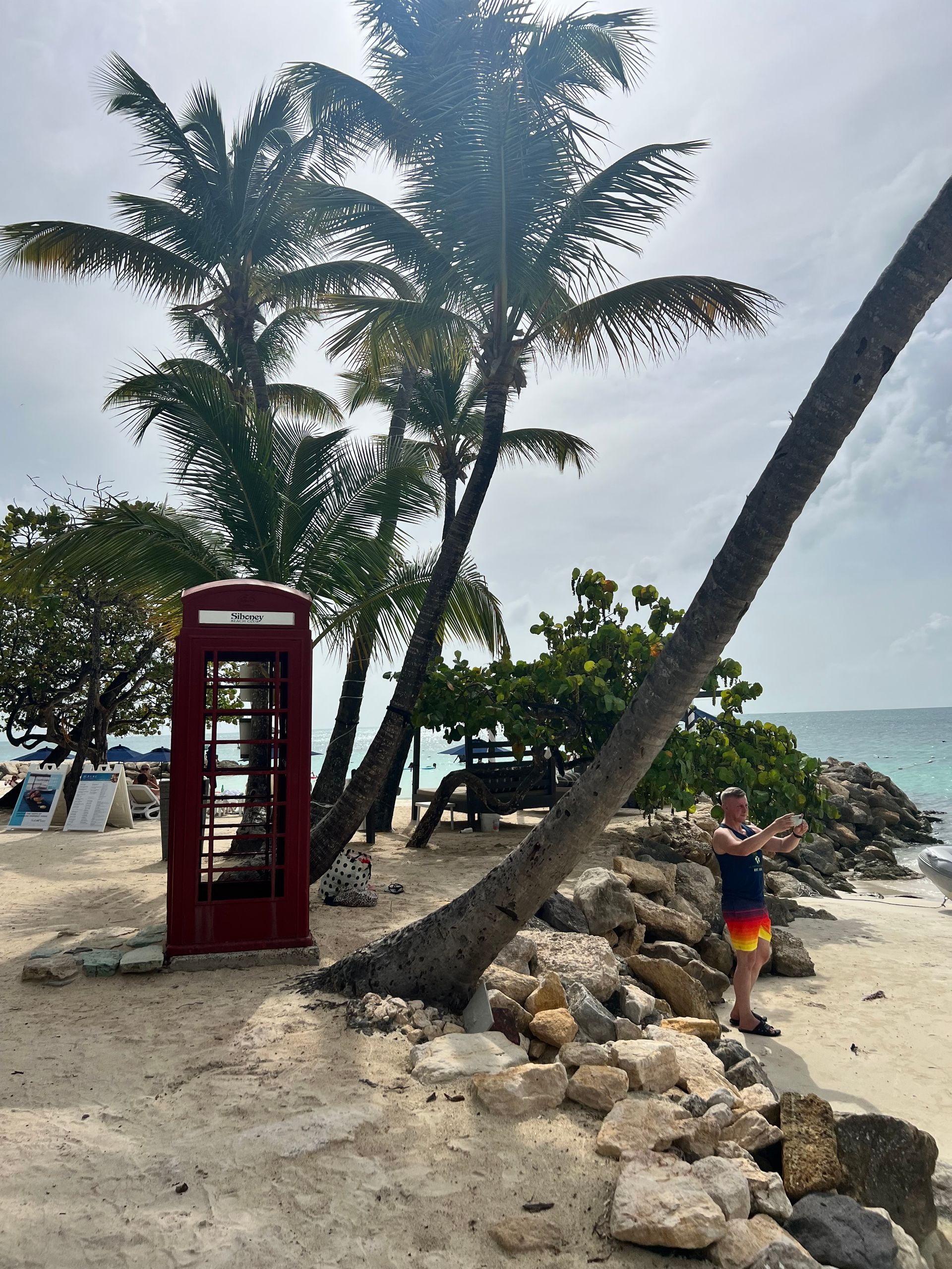 Person in colorful shorts poses near a red phone booth on a sandy beach with palm trees.