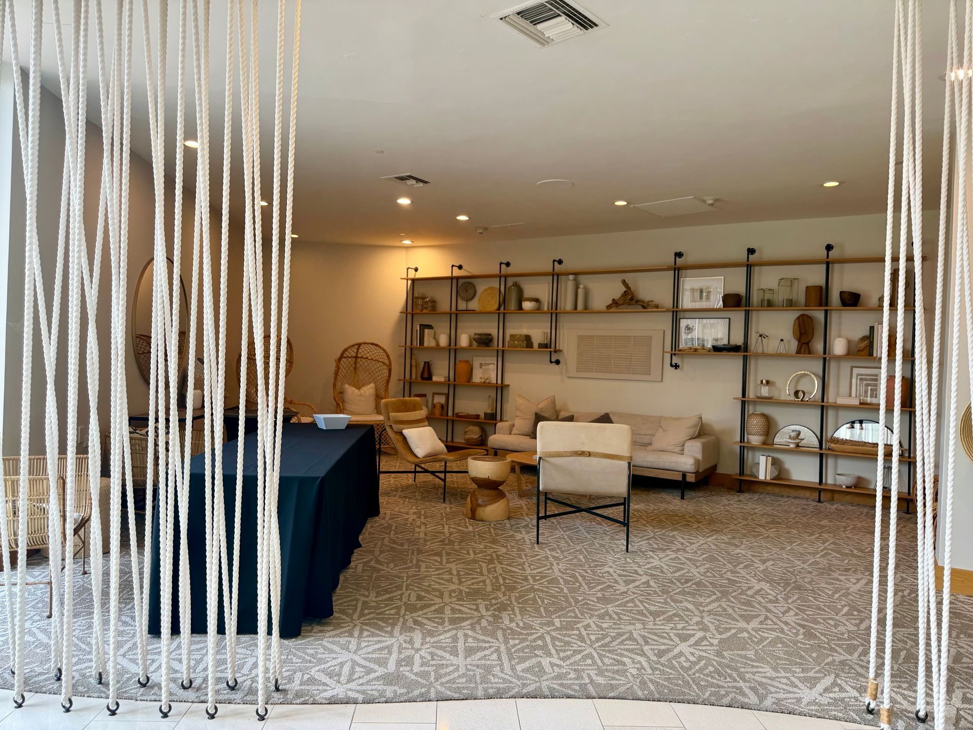 Modern lounge with shelving, beige chairs, and a woven floor rug behind a white string curtain
