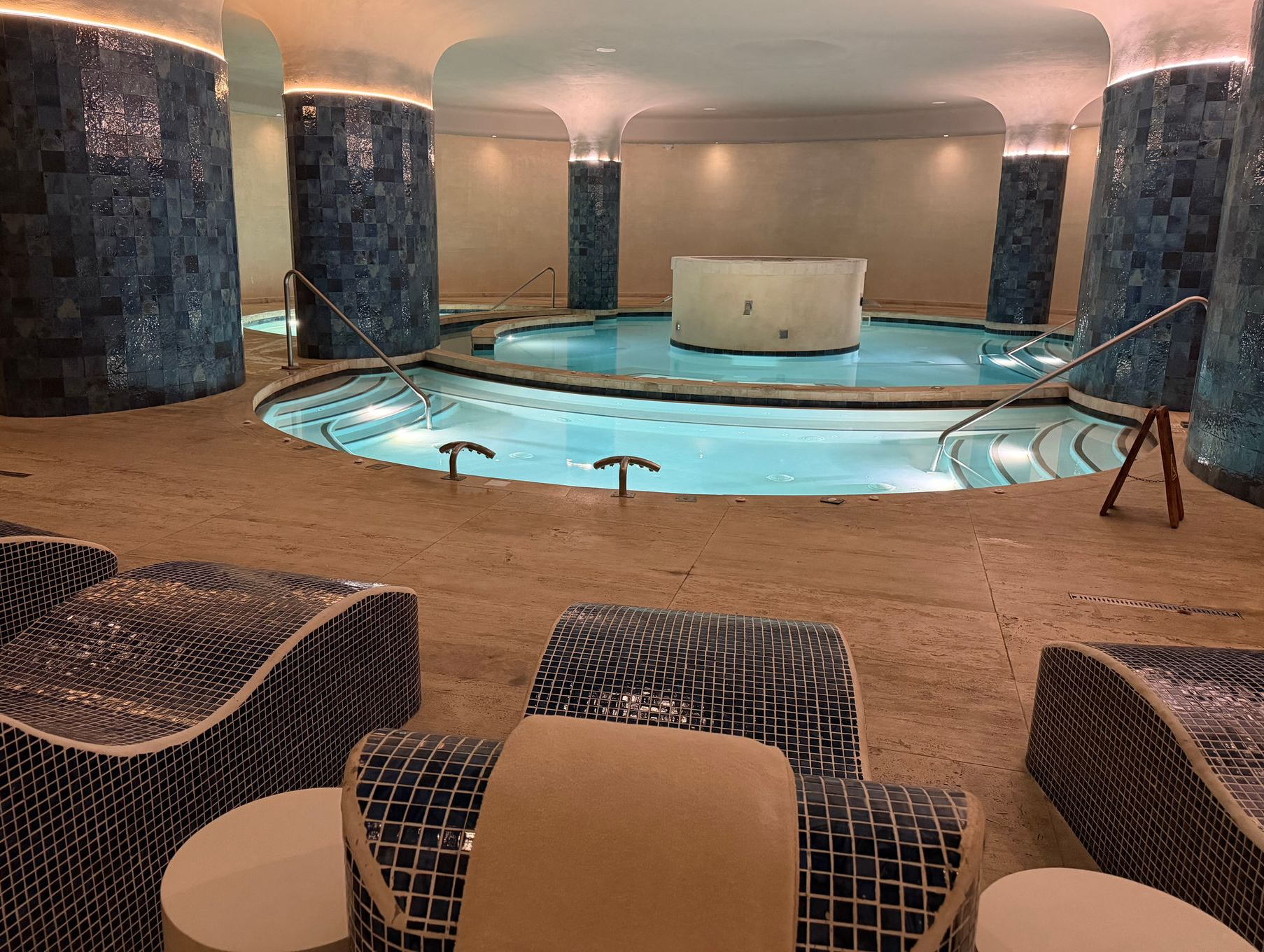 Indoor spa with a round pool, blue mosaic columns, and lounge chairs along the deck