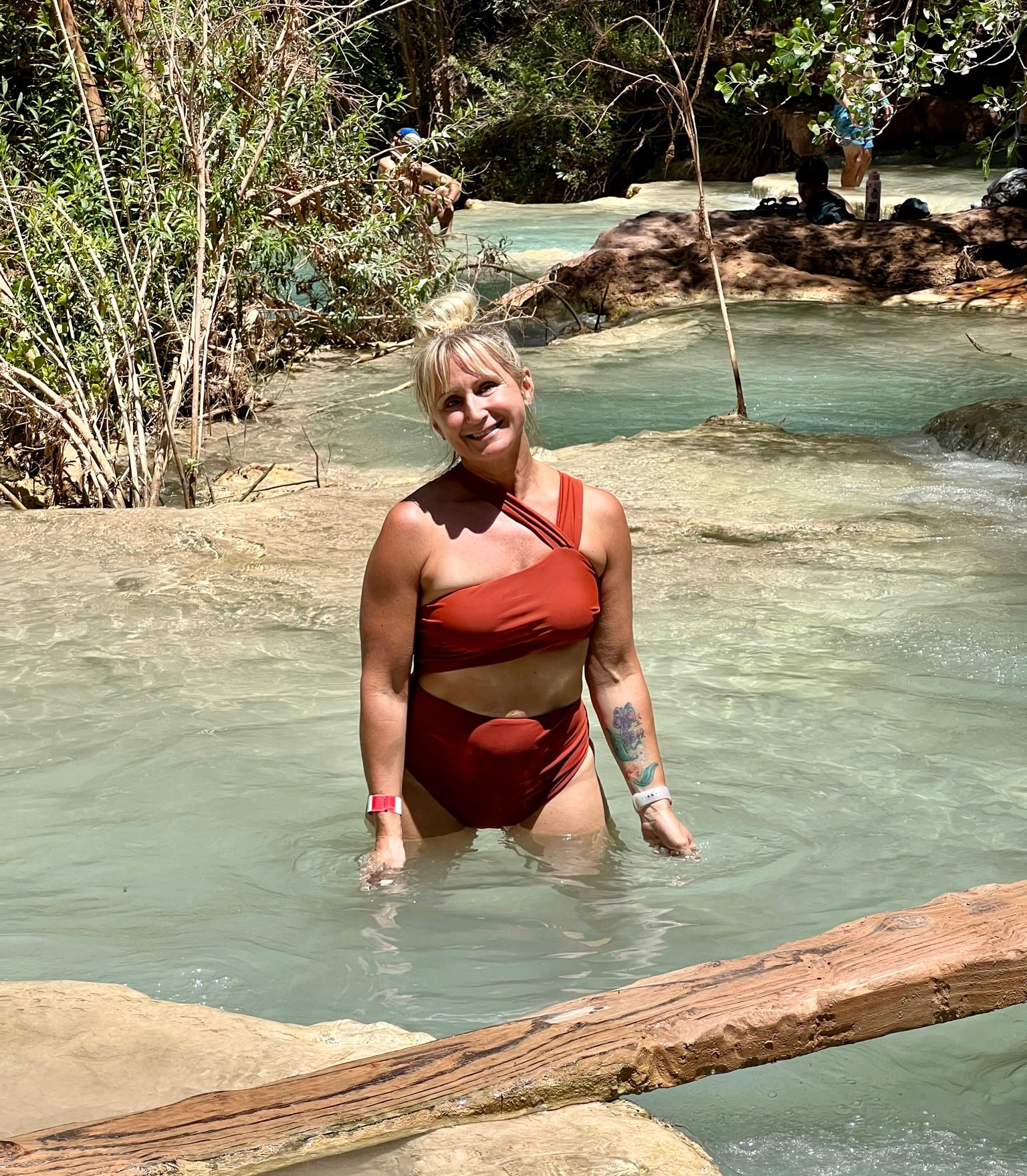 Person in a brown swimsuit standing in shallow turquoise water near a sandy stream.