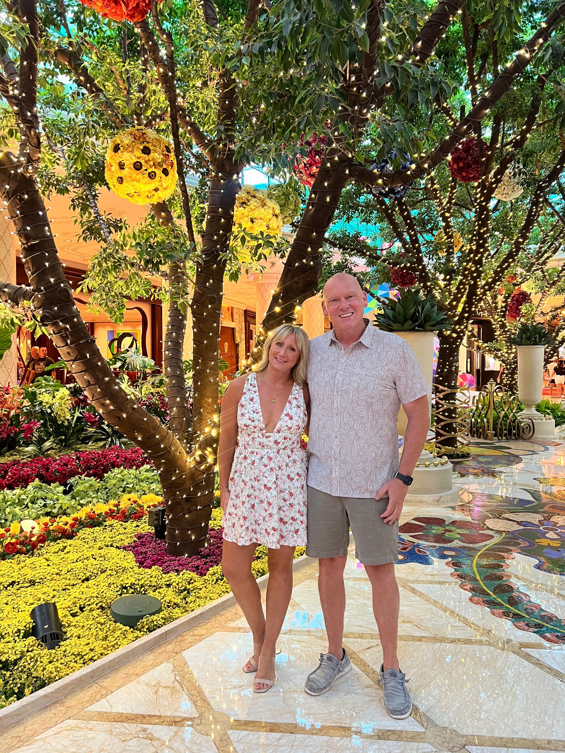 Couple poses under a tree with lights and floral decor. Woman in dress, man in patterned shirt, smiling. Las Vegas hotel