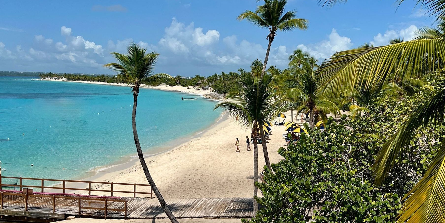Beach with turquoise water, white sand, palm trees, and people walking. Sunny day.