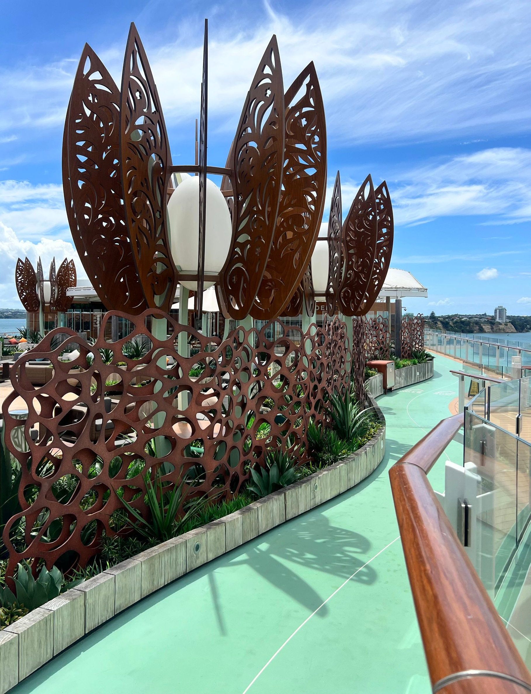 A green deck pathway on a cruise ship with decorative, leafy metal sculptures and blue sky.