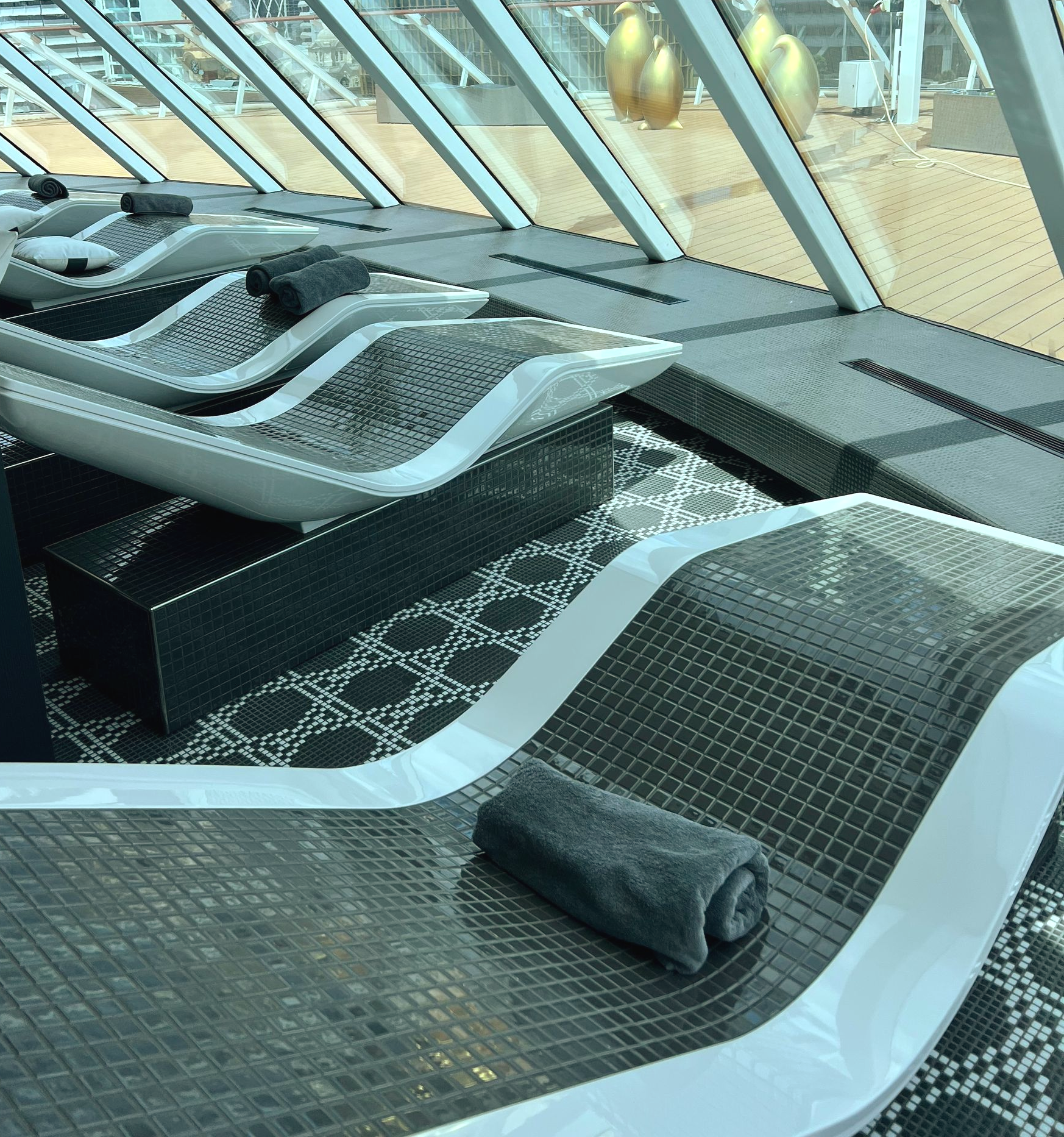 Lounge chairs with rolled towels, indoor spa area, angled glass walls on a cruise, geometric patterned floor.