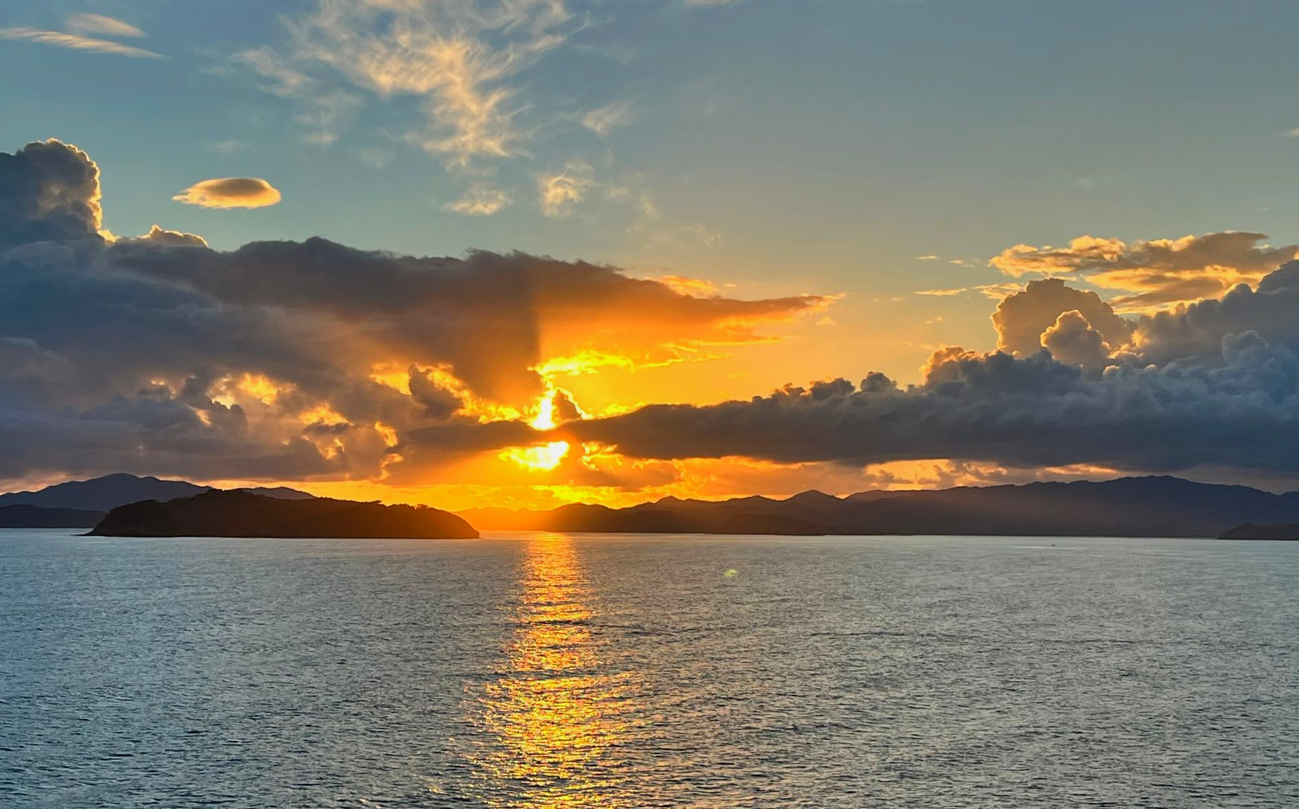 Sunset over water with sun breaking through clouds, island in the distance.