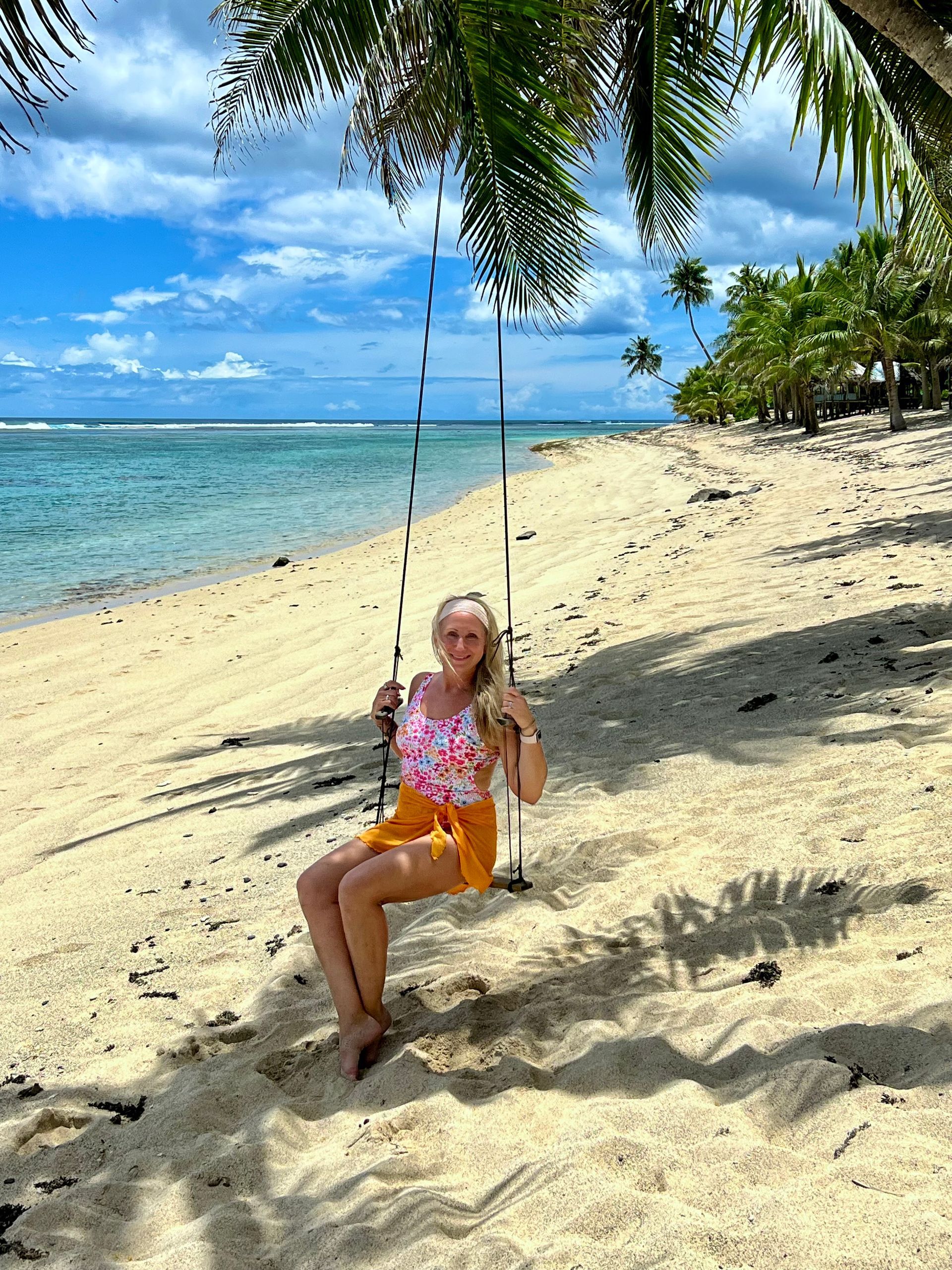 Woman on swing at beach, blonde hair, floral swimsuit, orange shorts, blue ocean, sunny day.