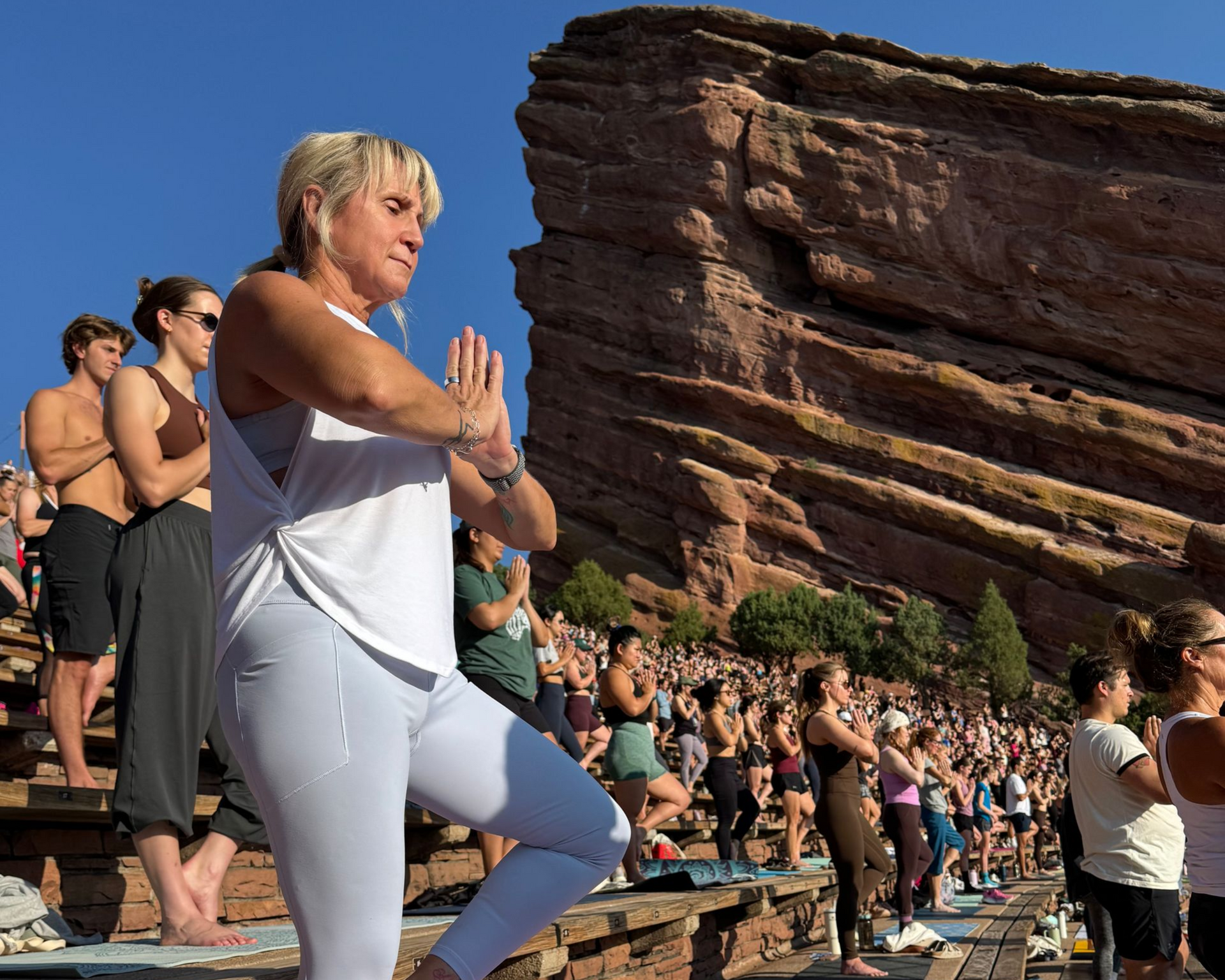 Woman practicing yoga on a mat outdoors at a rocky amphitheater, with a crowd seated behind her.