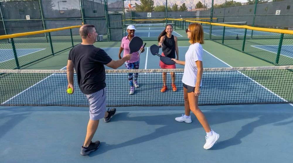 Four people stand on a pickleball court around the net, holding paddles and talking in casual sportswear.