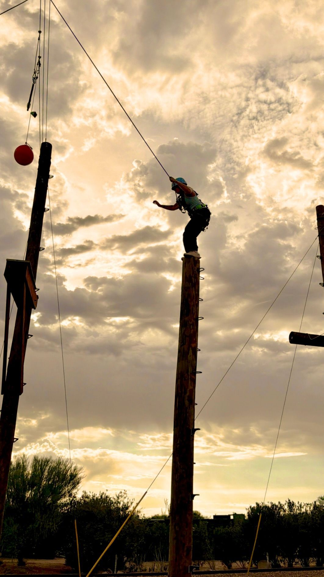 Woman climber on a tall wooden pole at an outdoor ropes course under a cloudy sky