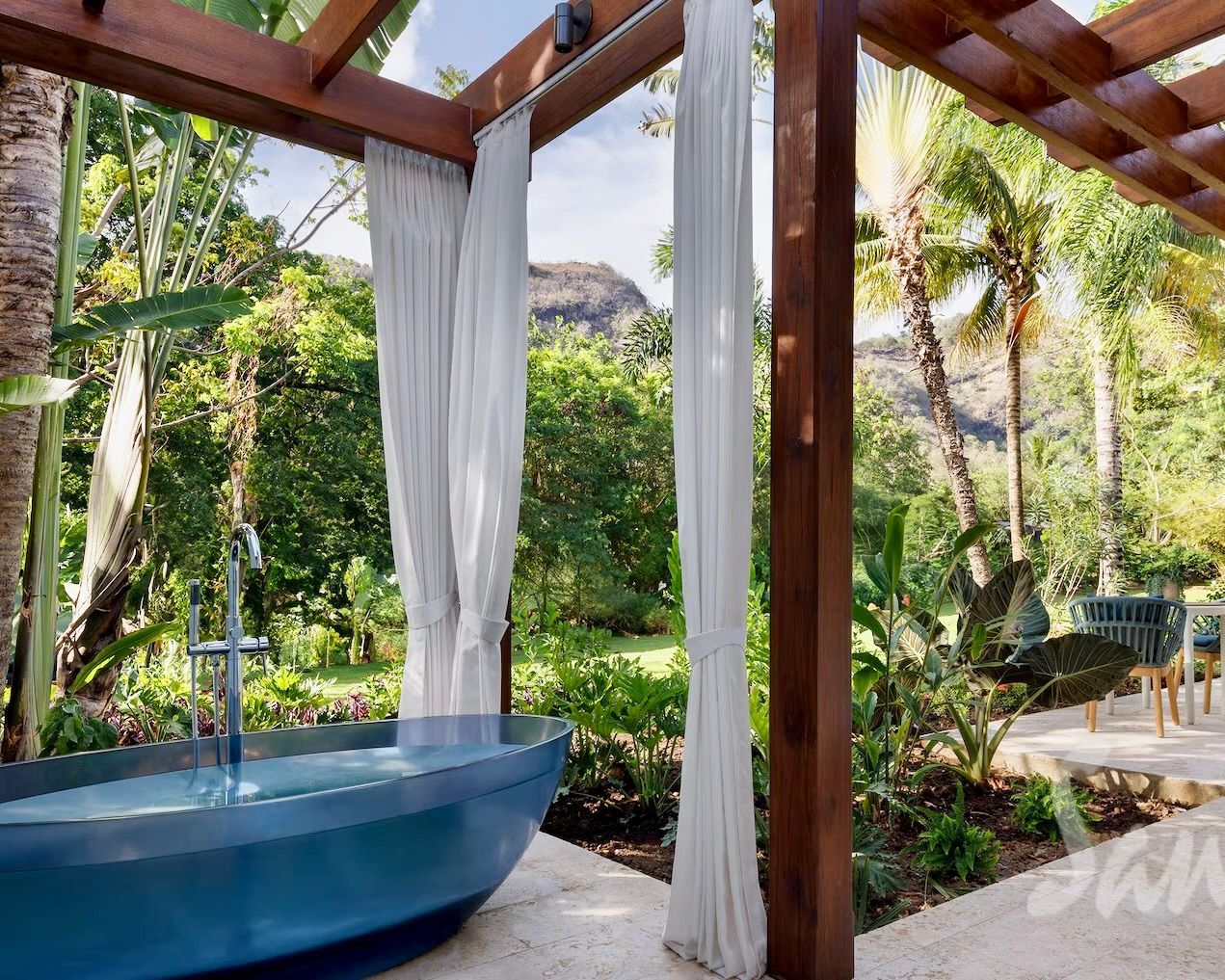Open-air patio with blue soaking tub, white curtains, and lush tropical garden view