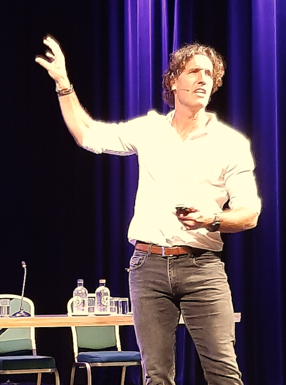 Martin Hersman als  op het podium, gebarend met één hand, met een afstandsbediening in de hand, voor paarse gordijnen en een tafel met waterflesjes.