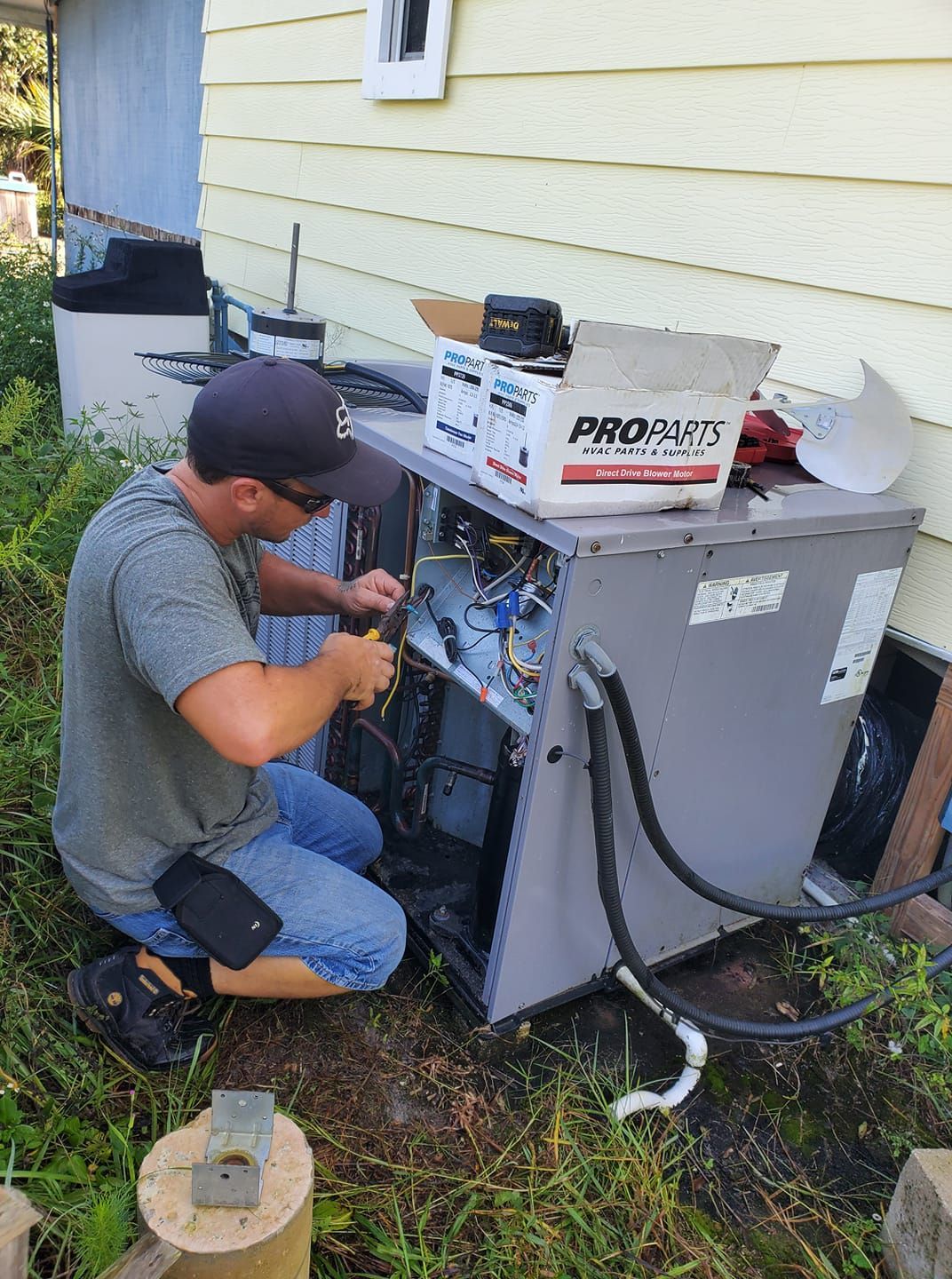 Man in baseball cap kneels, wiring an air conditioning unit. Exterior setting, beside a house, boxes nearby.