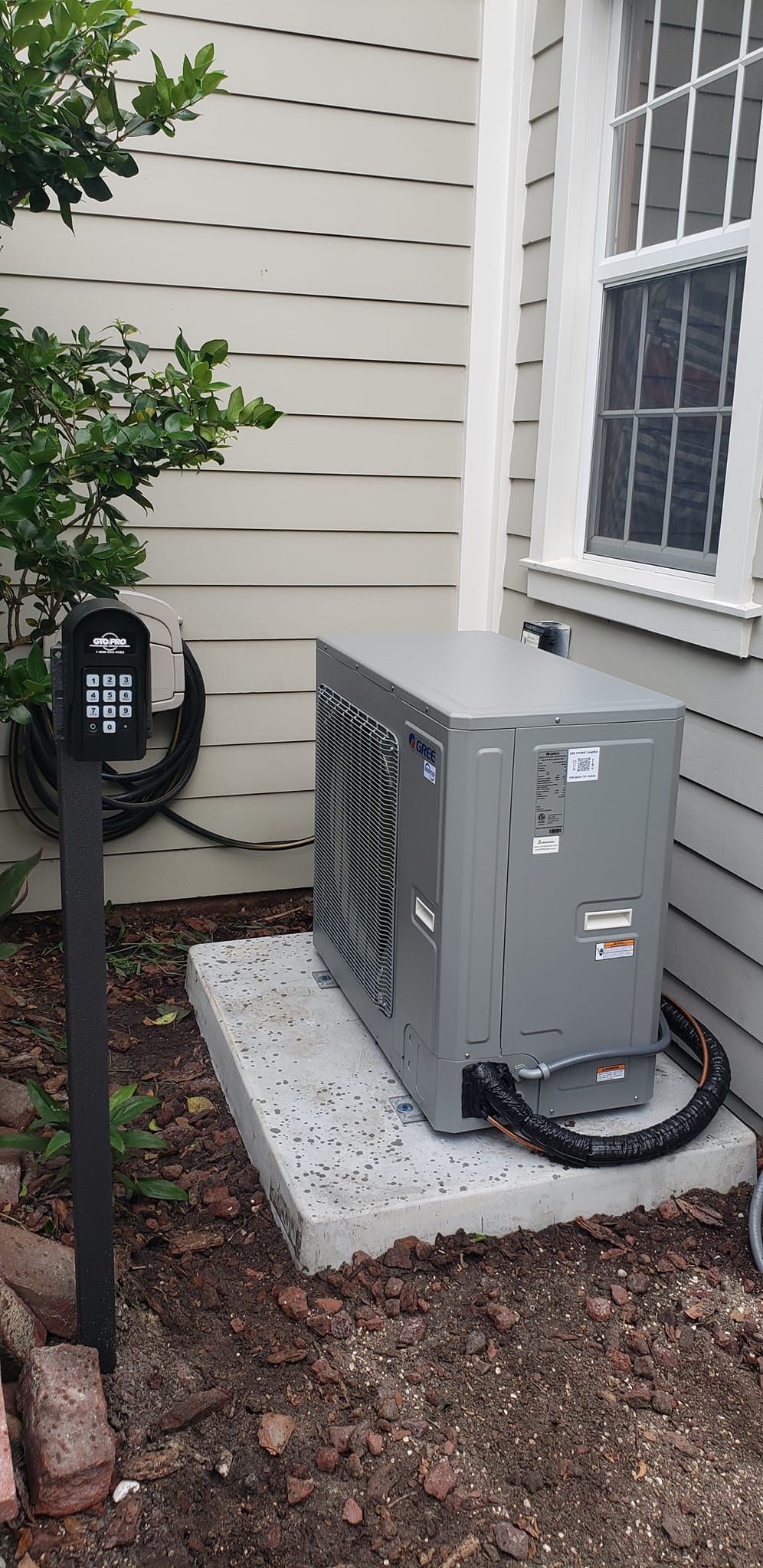 An air conditioning unit on a concrete pad next to a house with a window and a black keypad.