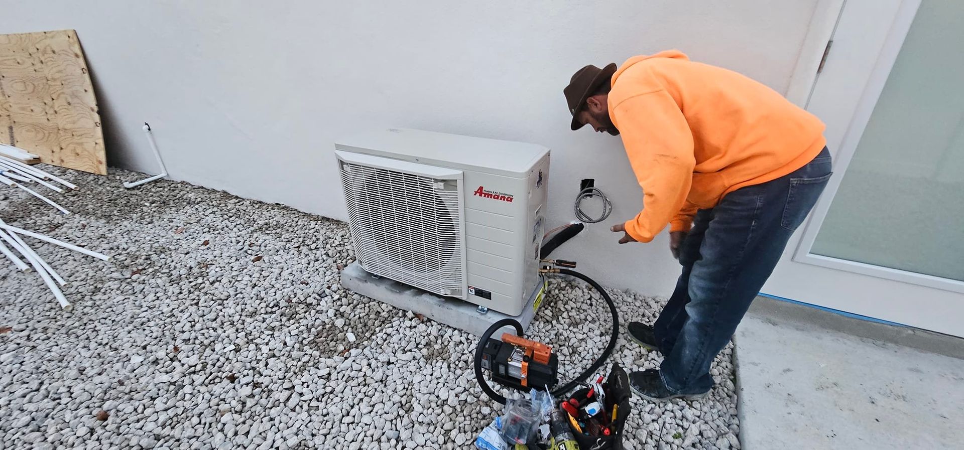 Man in orange shirt working on an AC unit outdoors next to a building on gravel.