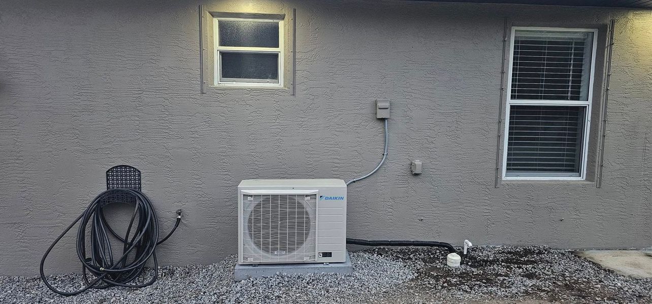 Exterior wall with two windows, a mounted hose, and an air conditioner unit. Gravel covers the ground.