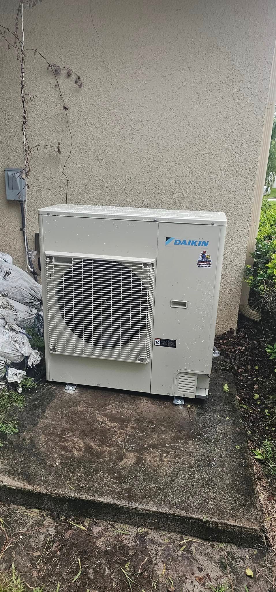 An air conditioner unit sits outside next to a concrete wall in the rain.