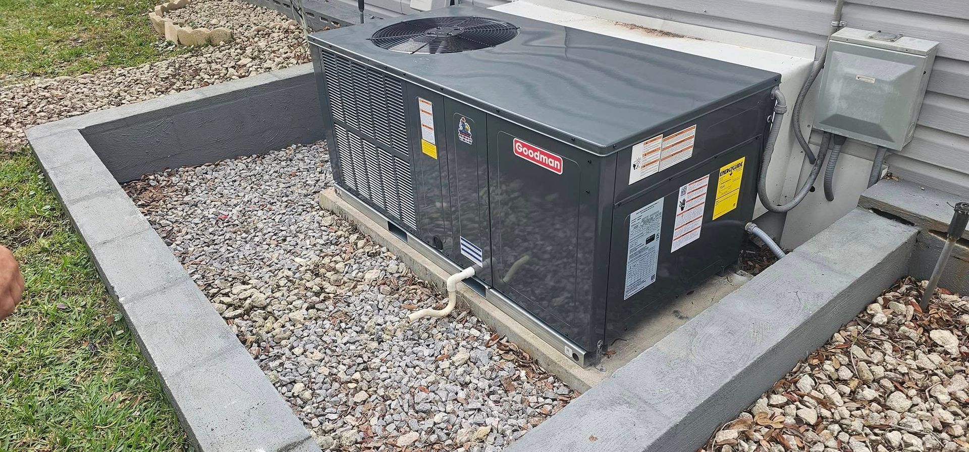 An air conditioner unit on a gravel base within a stone border. Next to a wall with electrical box.