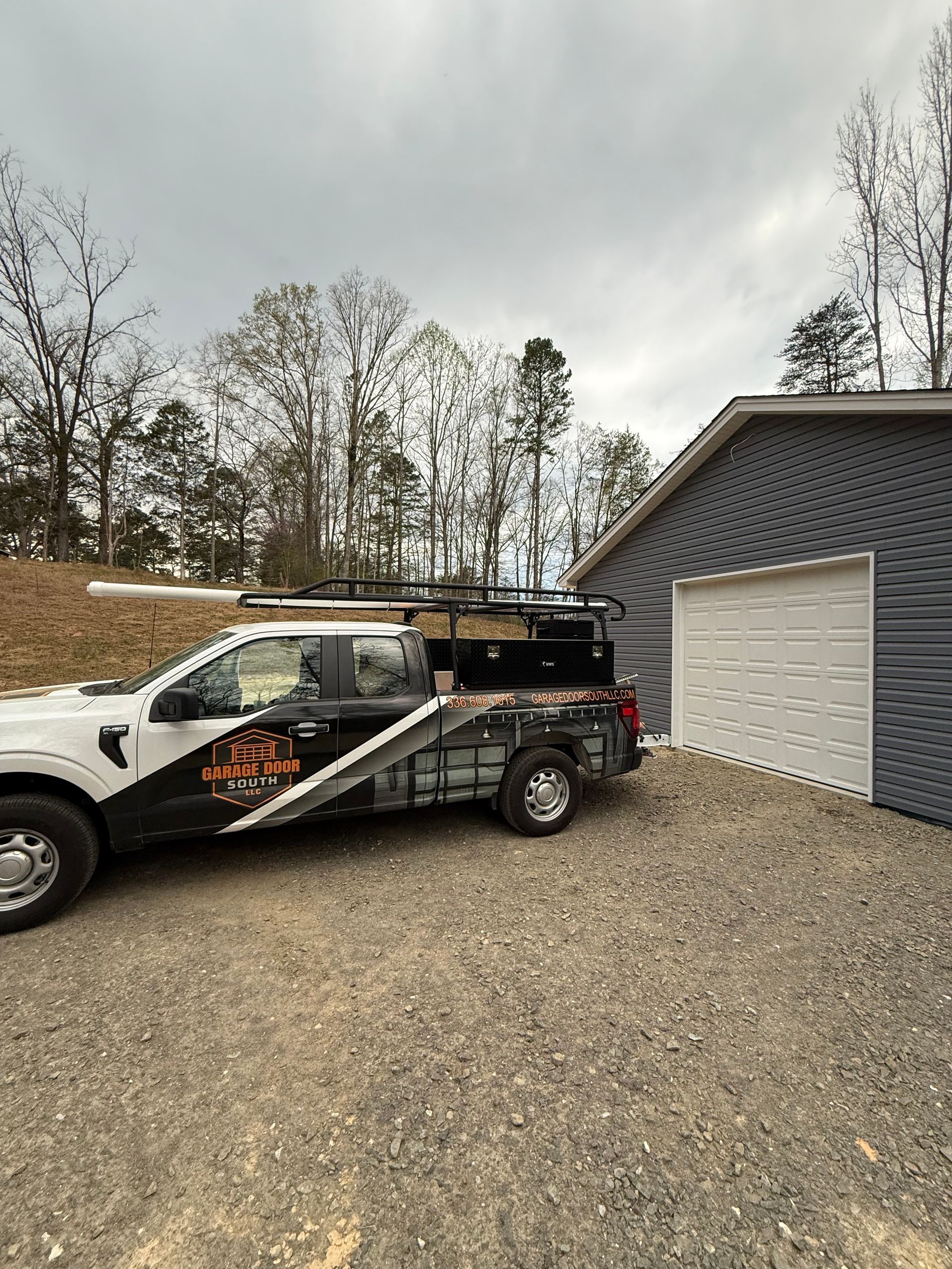 White work truck with a trailer parked on gravel beside a gray garage under cloudy skies.