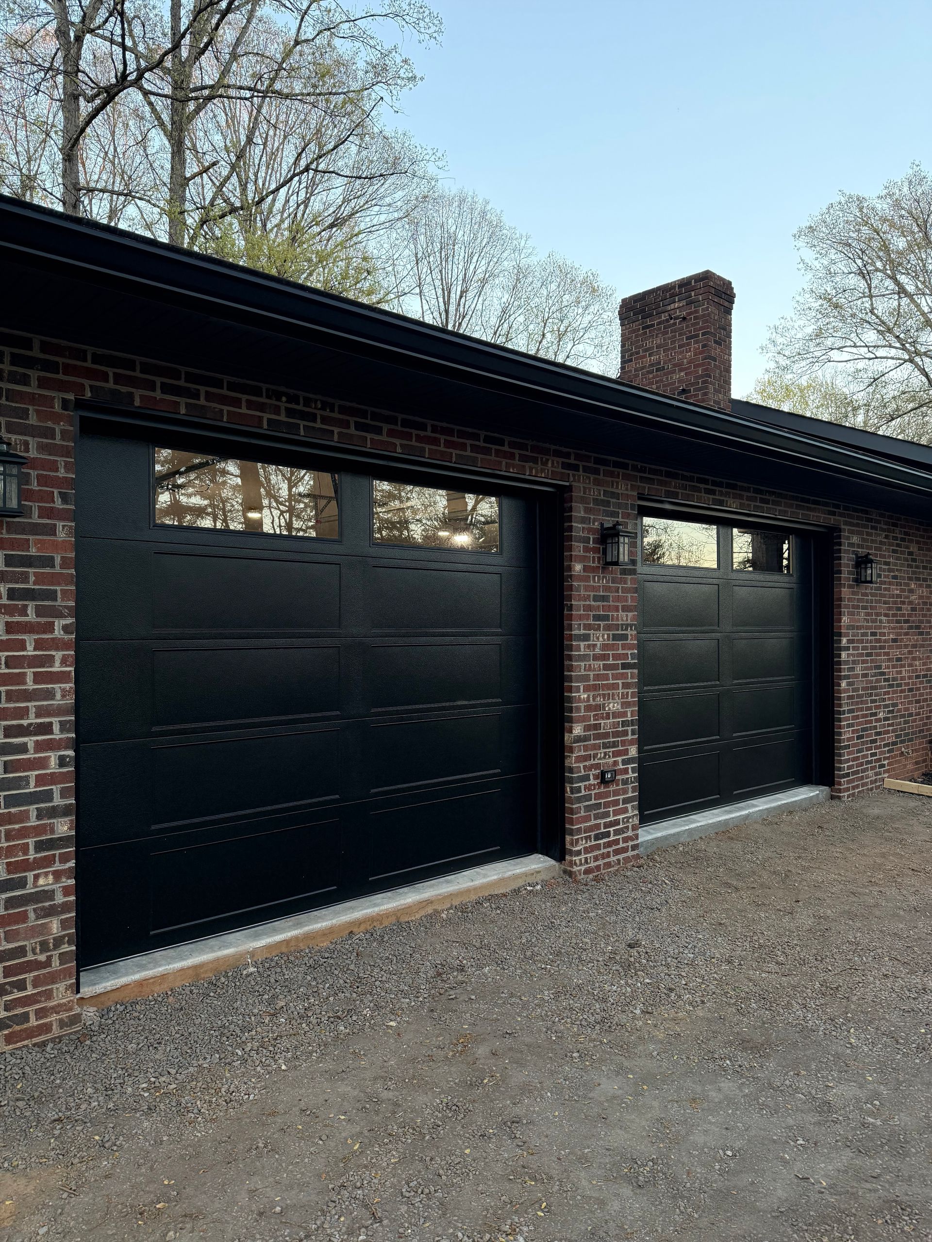 Black double garage doors on a brick house with gravel driveway and trees behind