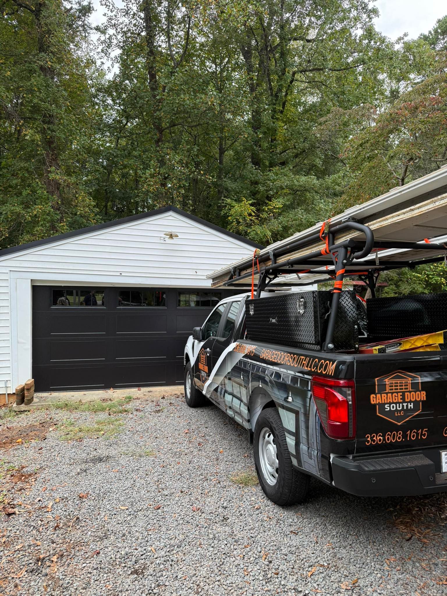 Black pickup truck with a ladder rack parked on gravel in front of a white garage and tall trees.