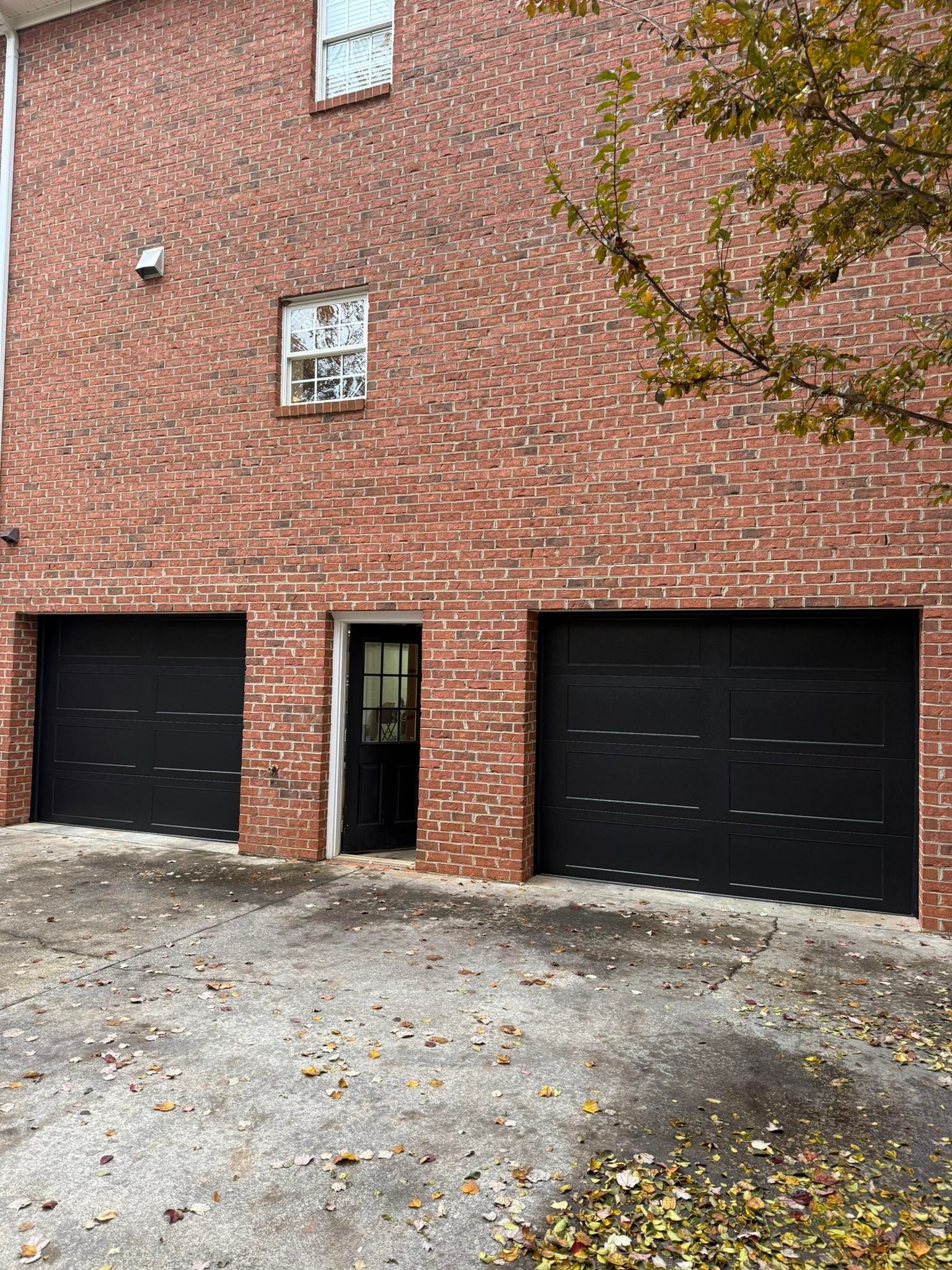 Brick apartment building with two black garage doors and a small entrance, partially covered by tree branches.
