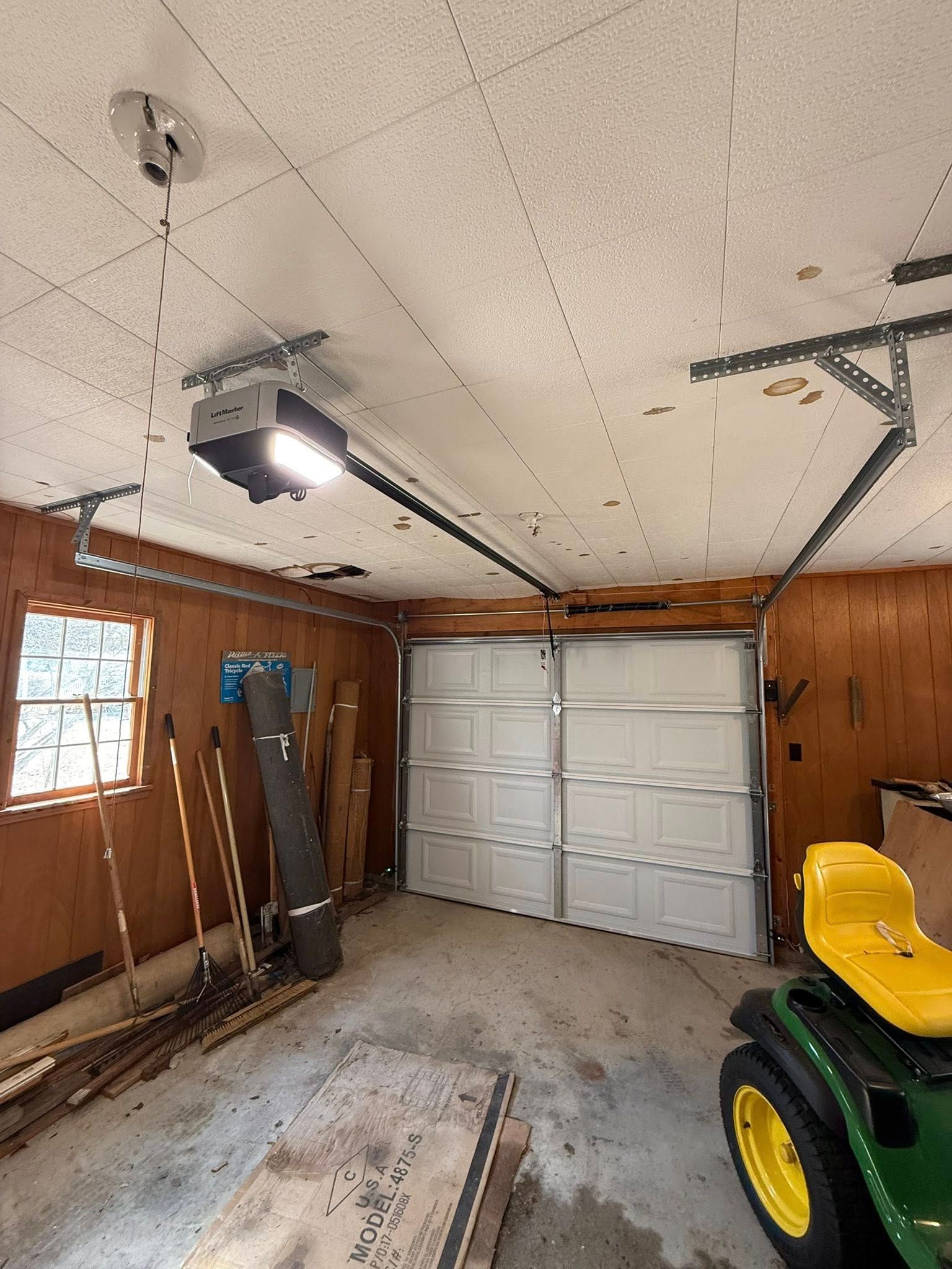 Garage interior with white roll-up door, wood-paneled walls, tools, and a yellow tractor.