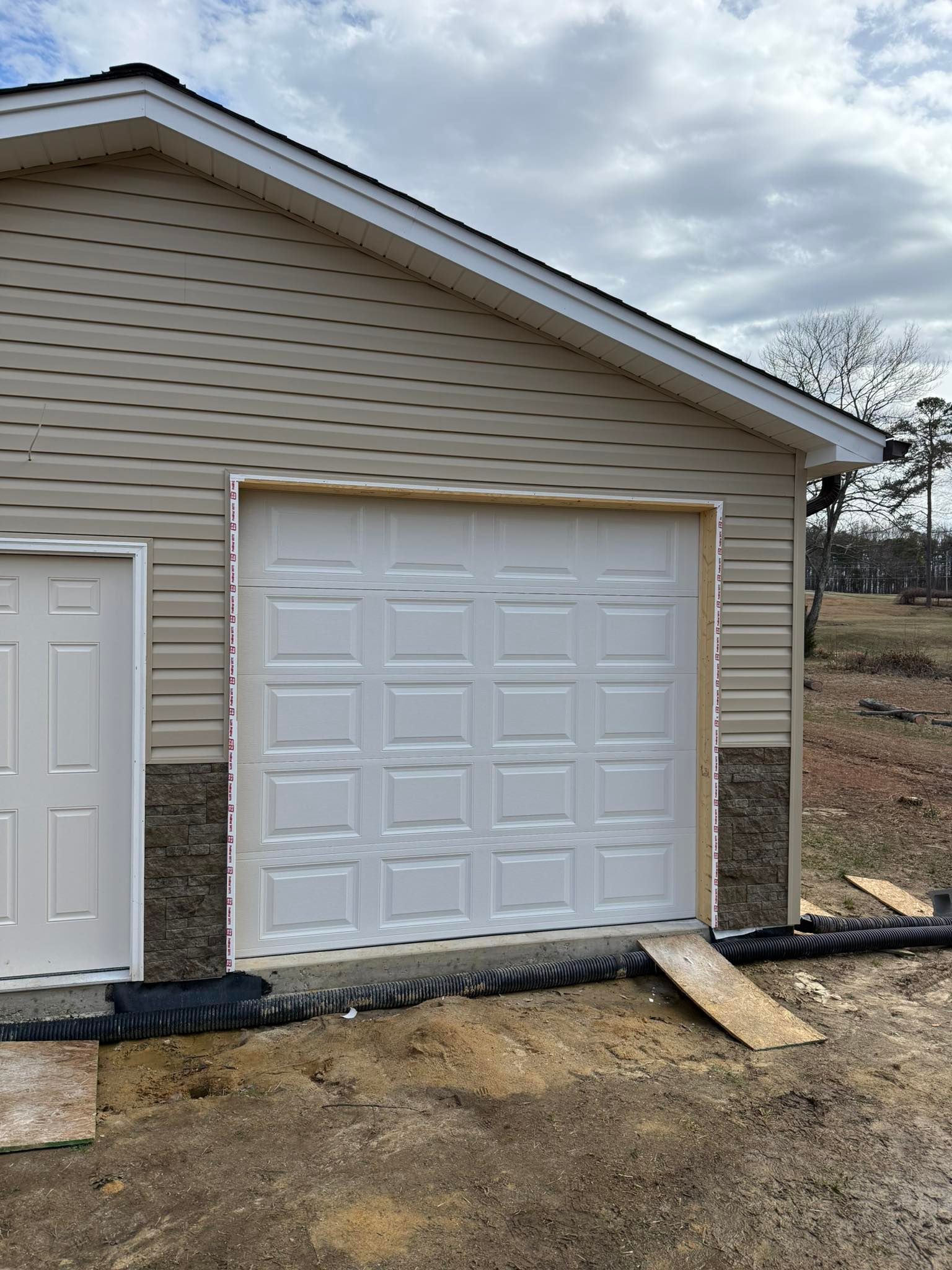 Detached garage with white door, beige siding, and a concrete ramp on a dirt lot under cloudy sky