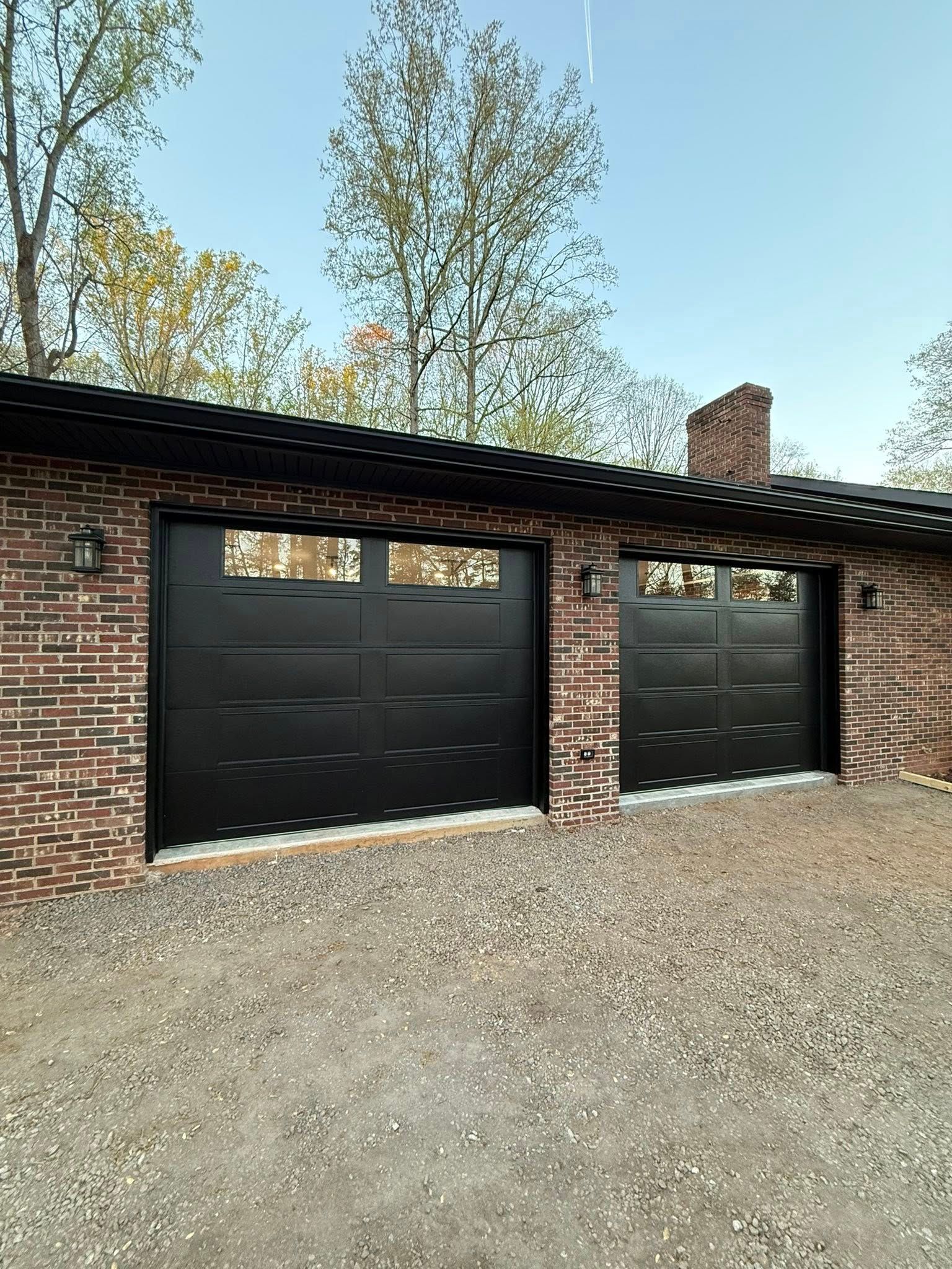 Brick garage with two black doors under a dark roof, set on a gravel driveway with trees behind.