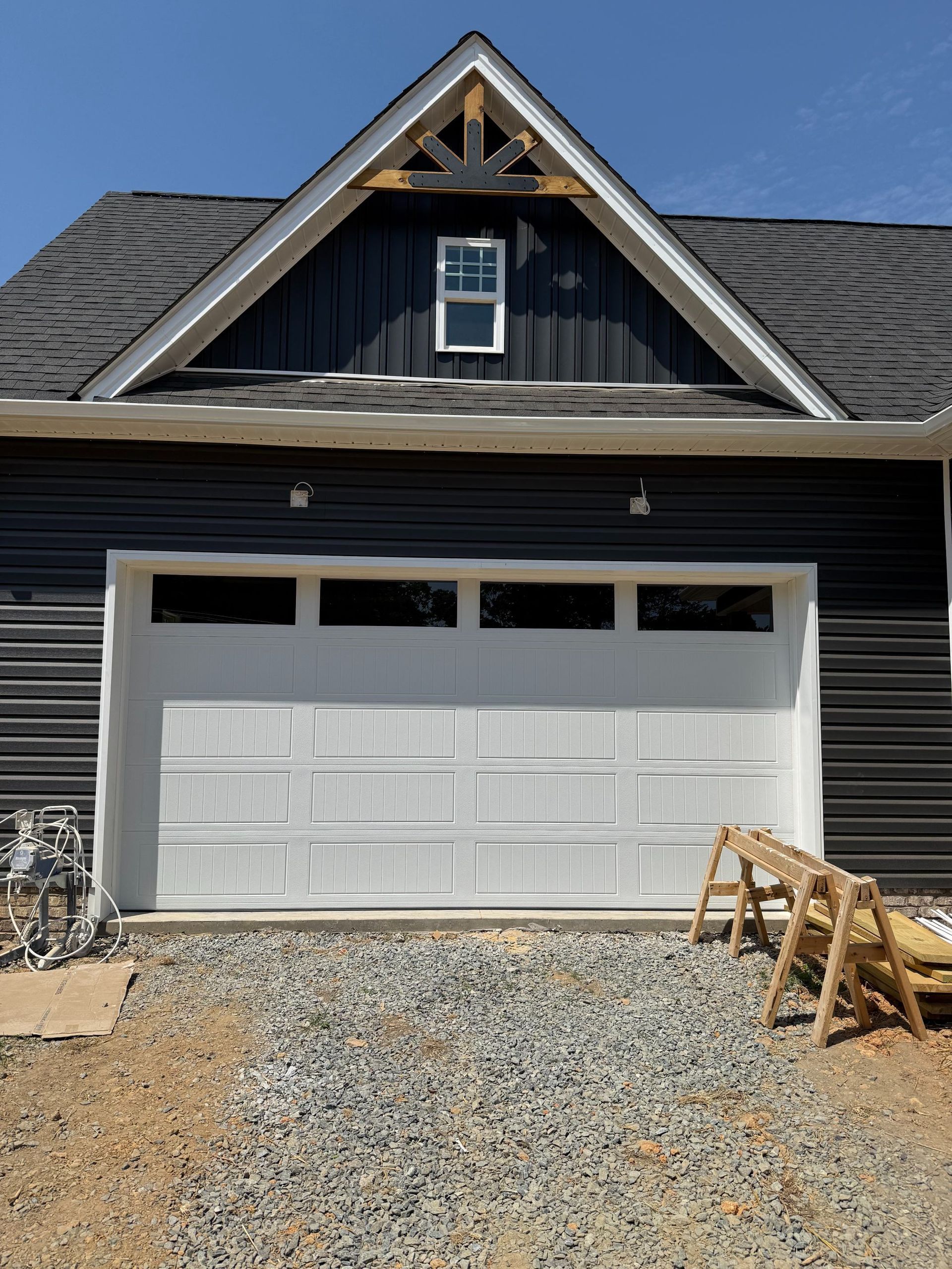 Black house exterior with a white garage door and gravel driveway, lumber stacked at right