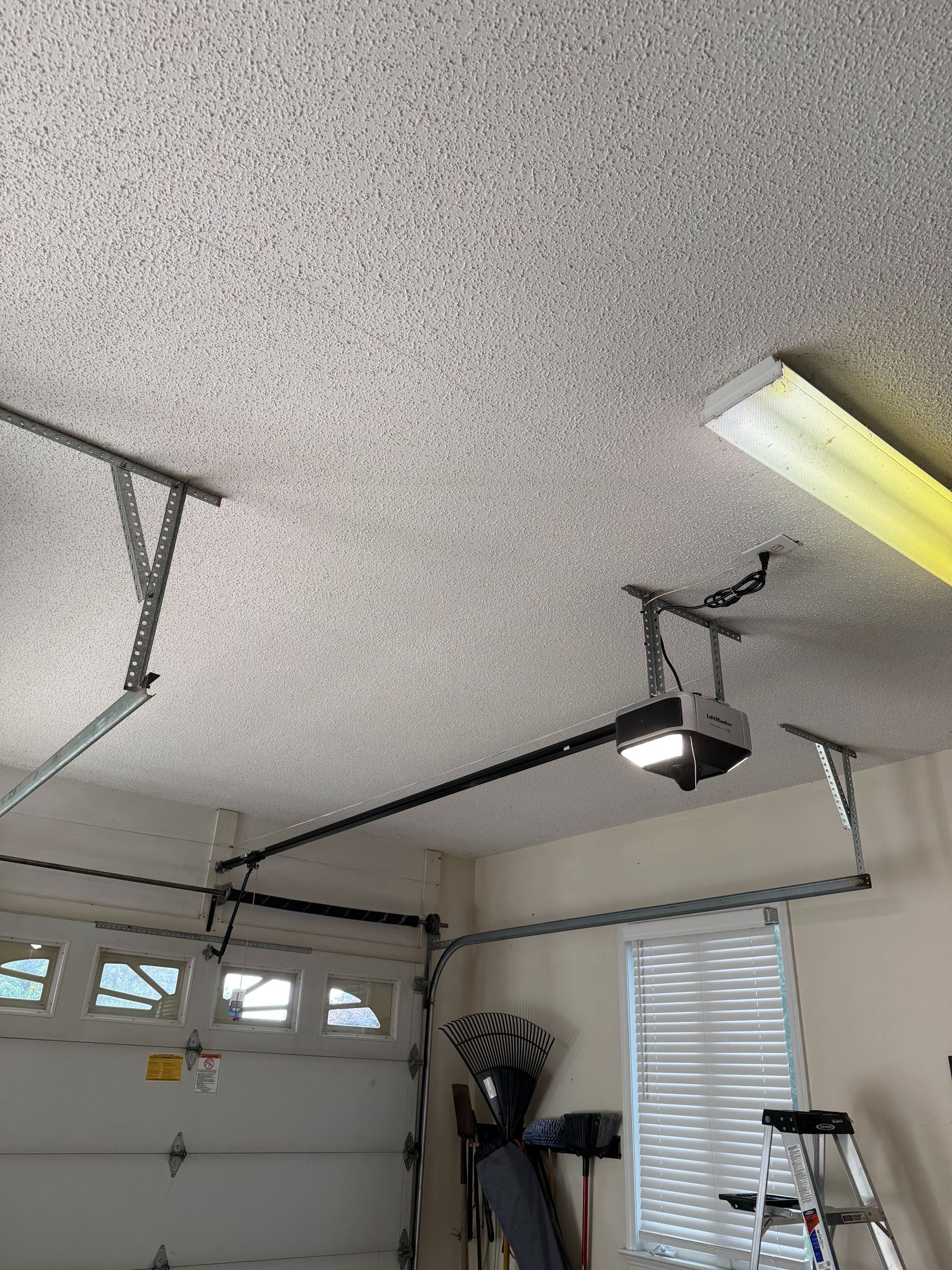 Garage interior with closed white door, ceiling opener, fluorescent light, and stored tools near a window.