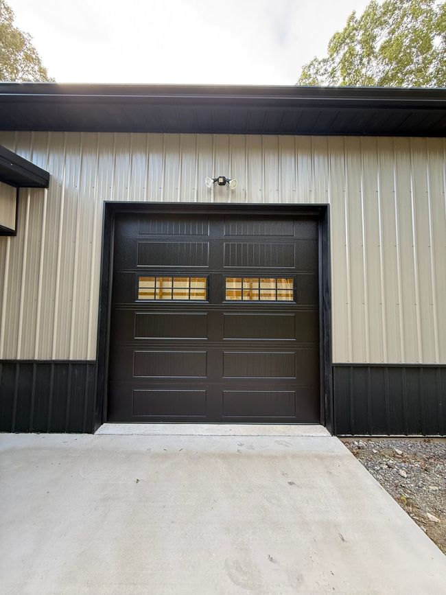 Brick garage with two black doors and a covered roof, set on a gravel driveway with trees behind it