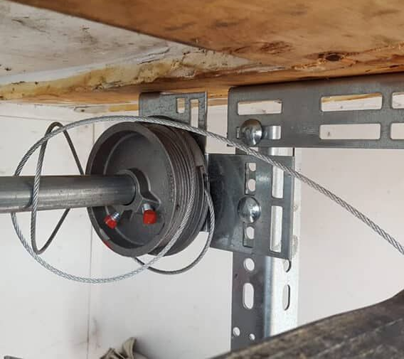 Close-up of a metal pulley and cable mounted to a gray bracket under a wooden shelf.