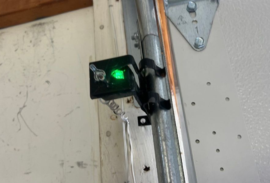 Green LED sensor mounted on a garage door track beside a white door panel