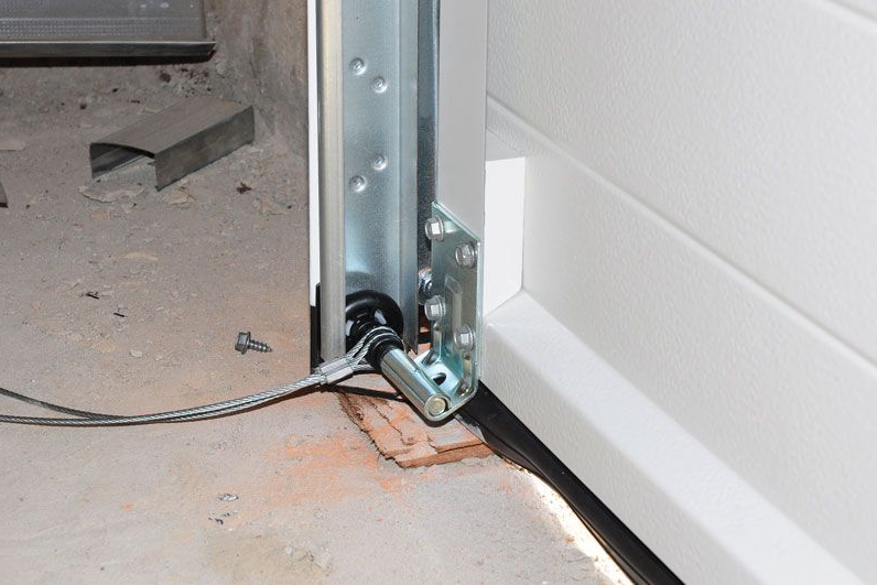 Close-up of a garage door track and bottom hinge beside a concrete floor, with a black cable nearby.