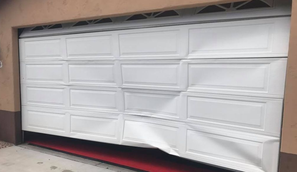 White garage door with visible dent and crease, viewed from the driveway.
