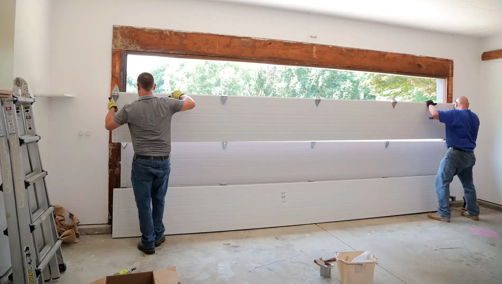 Two workers lifting a large white garage door into place in a bright room
