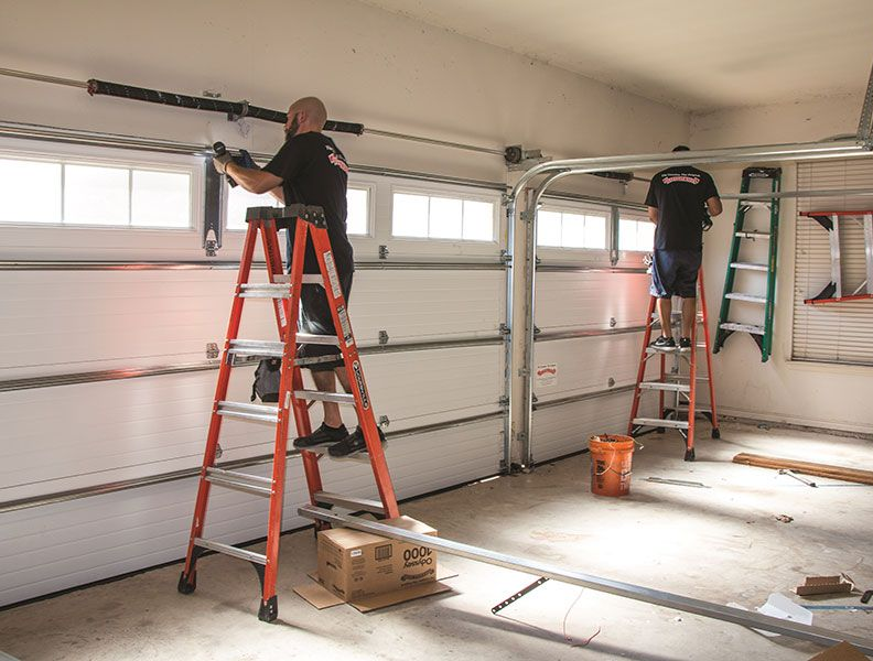 Two people on ladders installing a garage door opener in a bright garage
