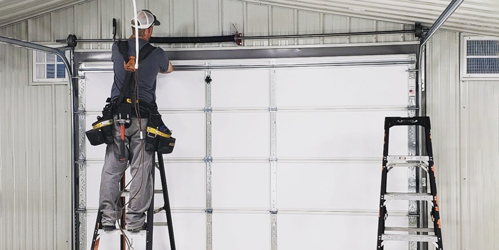 Worker on a ladder repairing a garage door inside a white industrial building