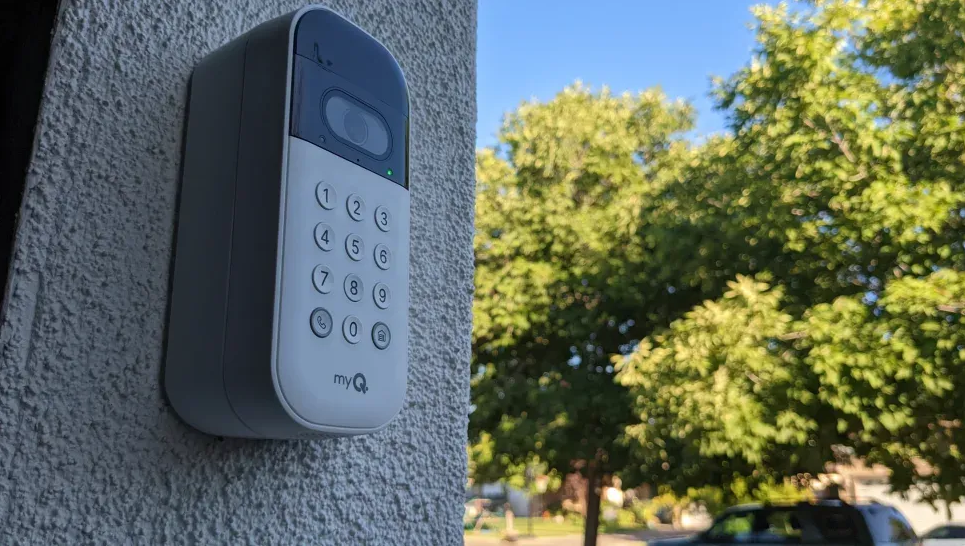 Gray keypad access intercom mounted on a stucco wall outdoors, with trees in the background