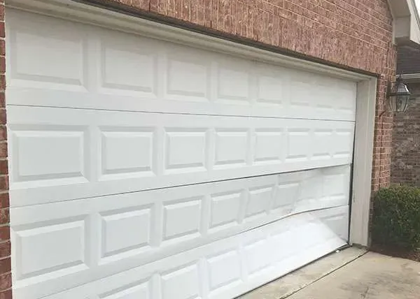 White garage door bent inward at the bottom, attached to a brick house driveway.