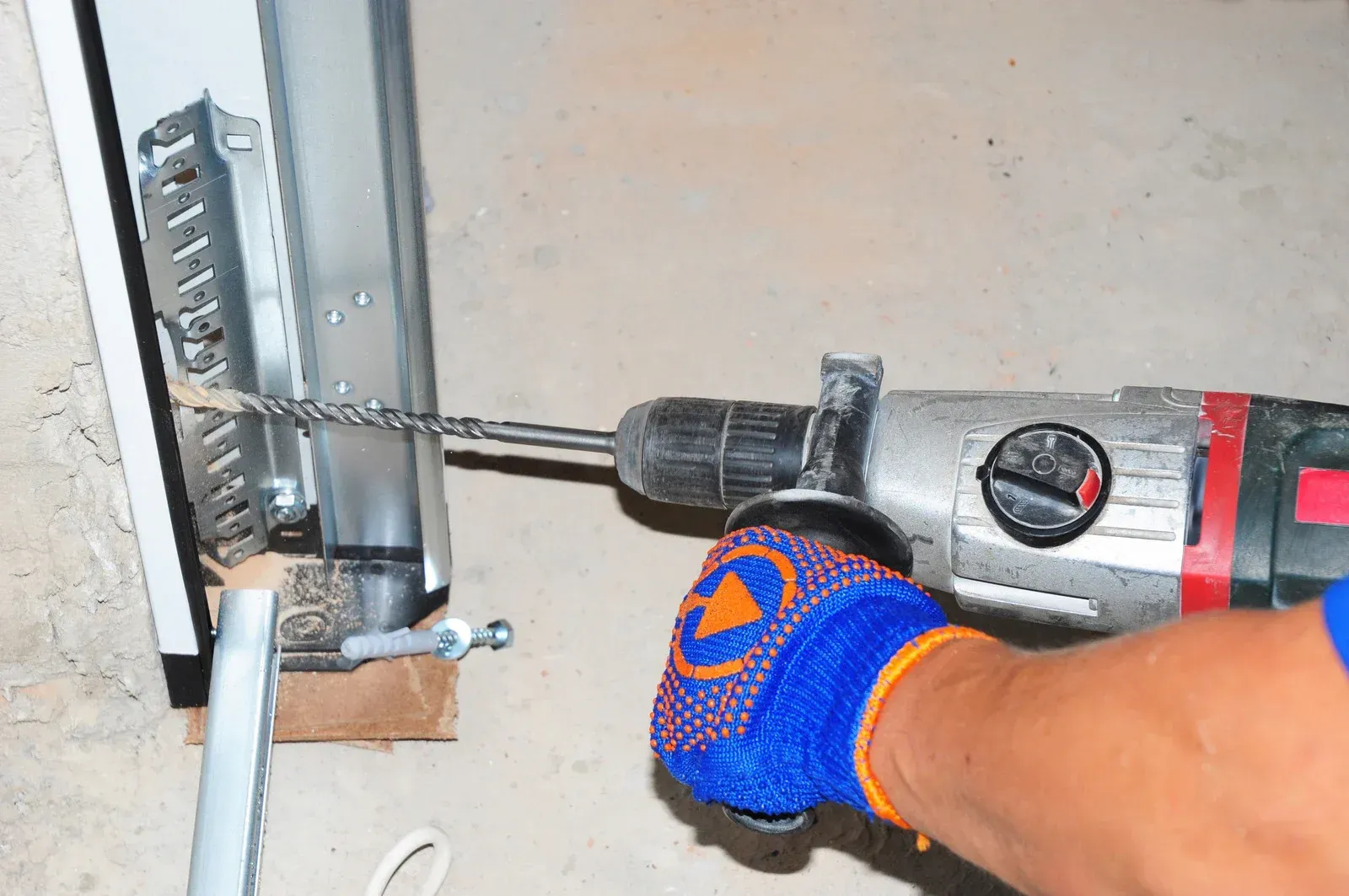 Gloved hand using a red-and-black drill to install a metal latch on a door frame