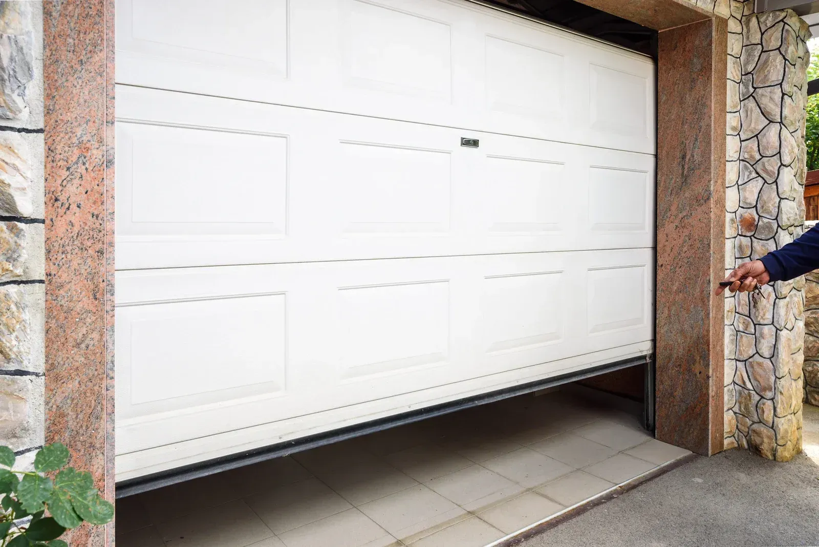 White garage door partially open at a stone house driveway, with a hand reaching from the right side
