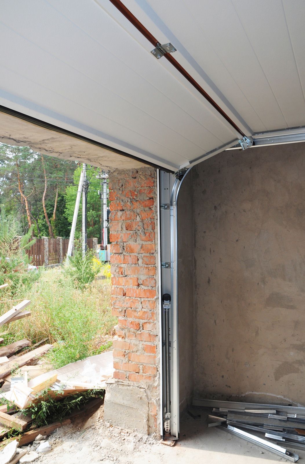 Open garage with metal door track, brick wall, and unfinished interior near a garden path