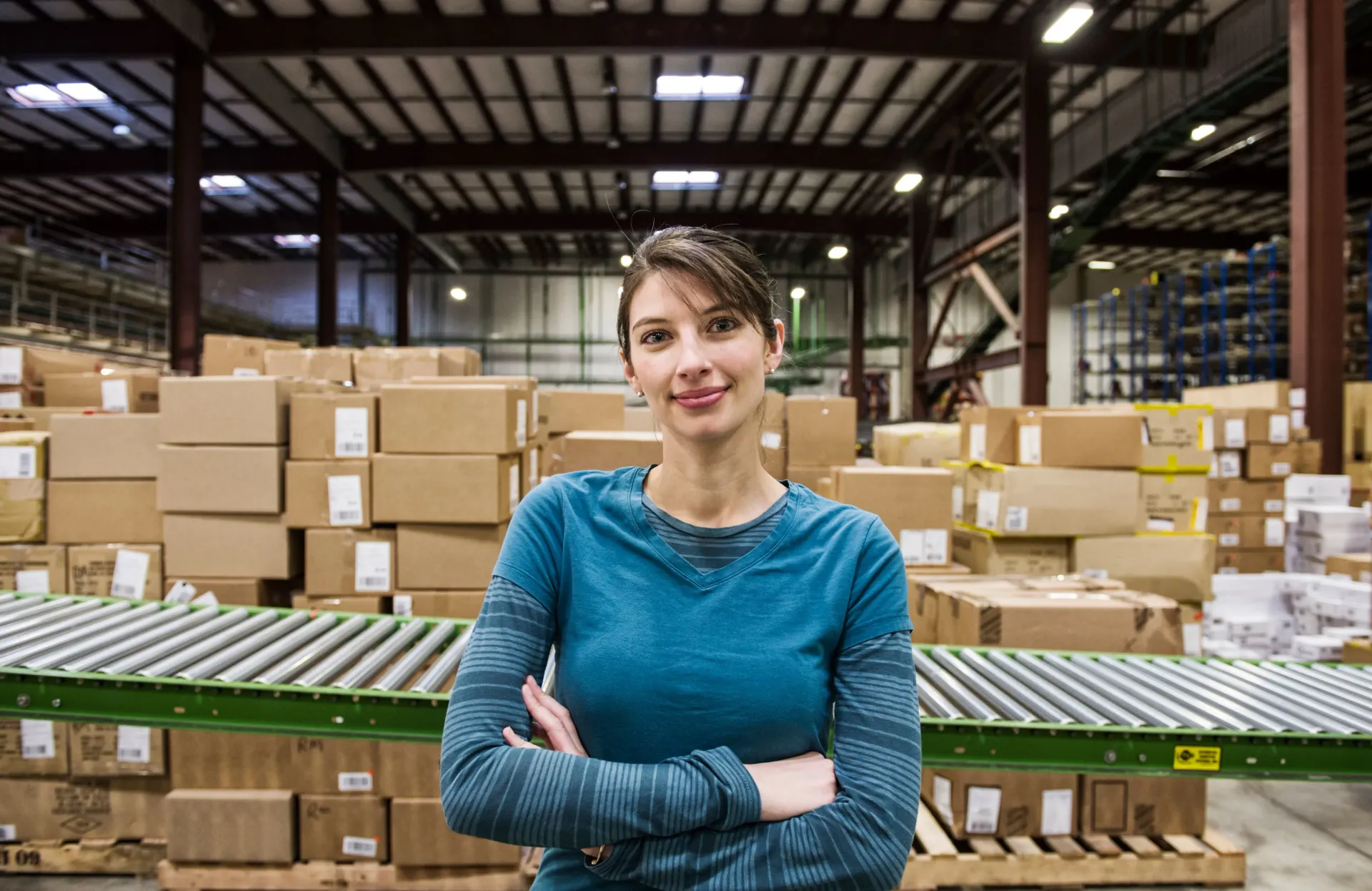 Woman standing in a warehouse with stacked boxes and conveyor belts, arms crossed, wearing a blue shirt