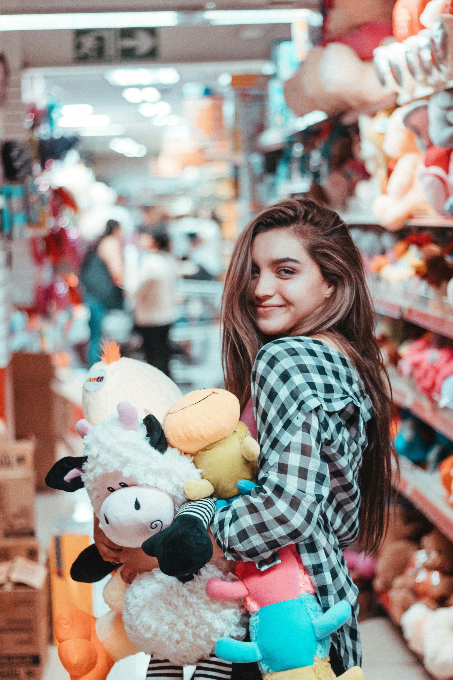 Mulher de camisa xadrez segurando brinquedos de pelúcia, sorrindo no corredor de uma loja de brinquedos.