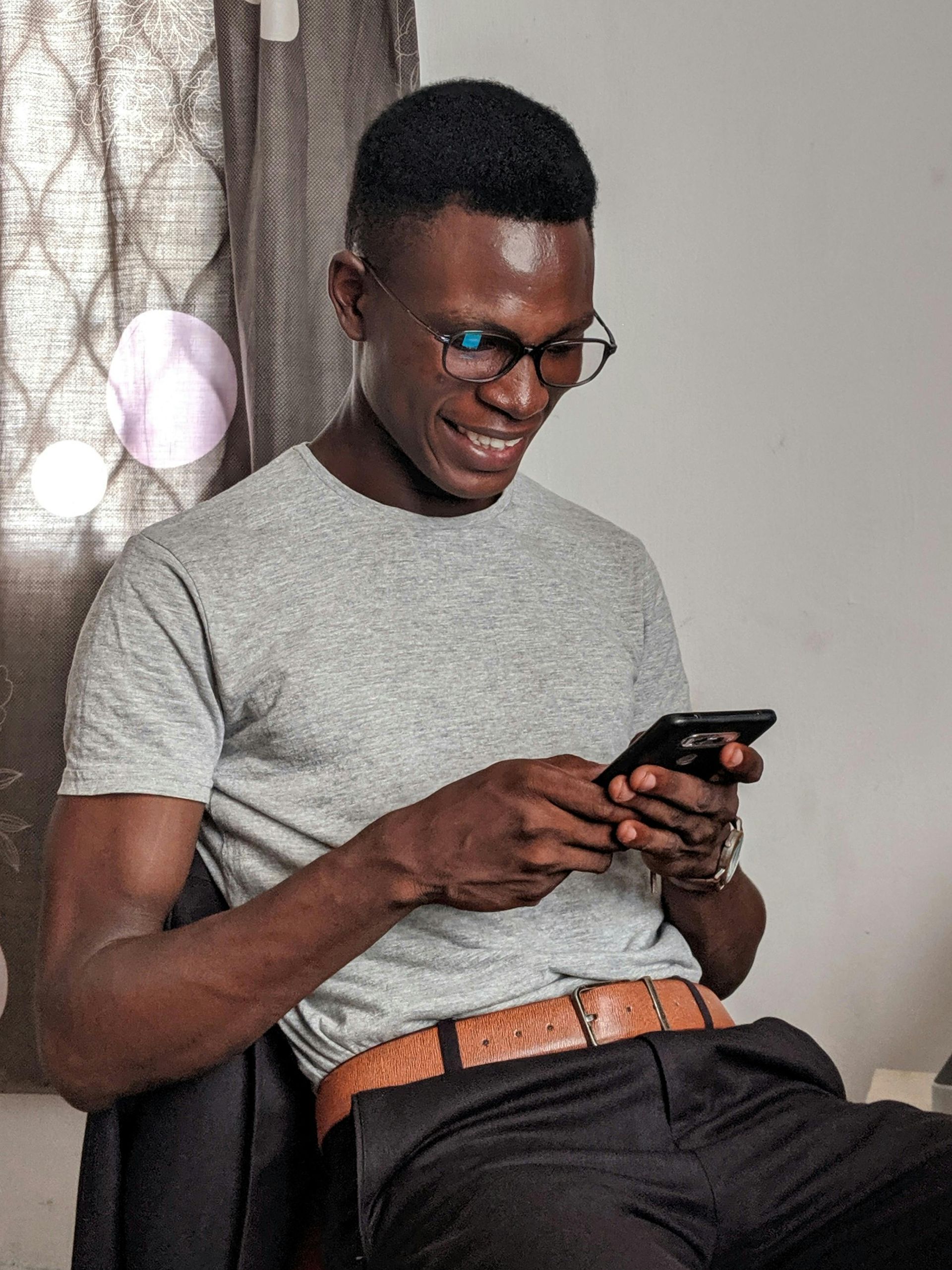 Um homem negro sorridente, usando óculos, envia mensagens de texto em seu telefone, sentado em uma sala com uma cortina.