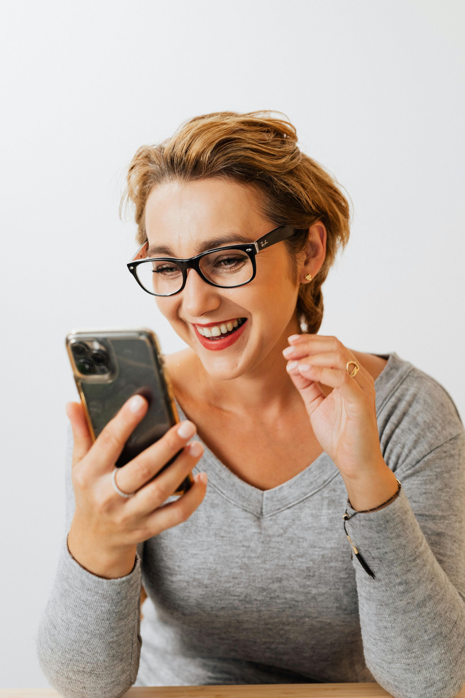 Mulher de óculos ri enquanto olha para o celular. Camisa cinza, cabelos claros, fundo branco.