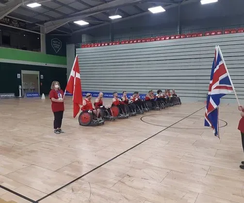 Kørestolsrugbyhold fra Danmark og Storbritannien stiller sig op til en begivenhed med flag på en indendørsbane.