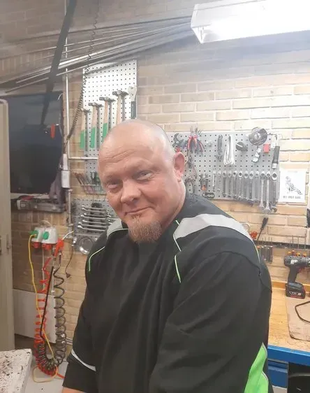 Mand med fipskæg i værksted, smilende, med værktøj på væggen bag sig.